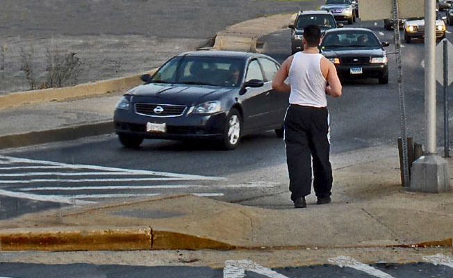 Photo shows a person standing on an island waiting to cross one lane of right-turning traffic.  A crosswalk is painted from the island across one lane to the sidewalk on the corner.  A long line of cars is approaching the crosswalk, and there is no traffic signal for that traffic.